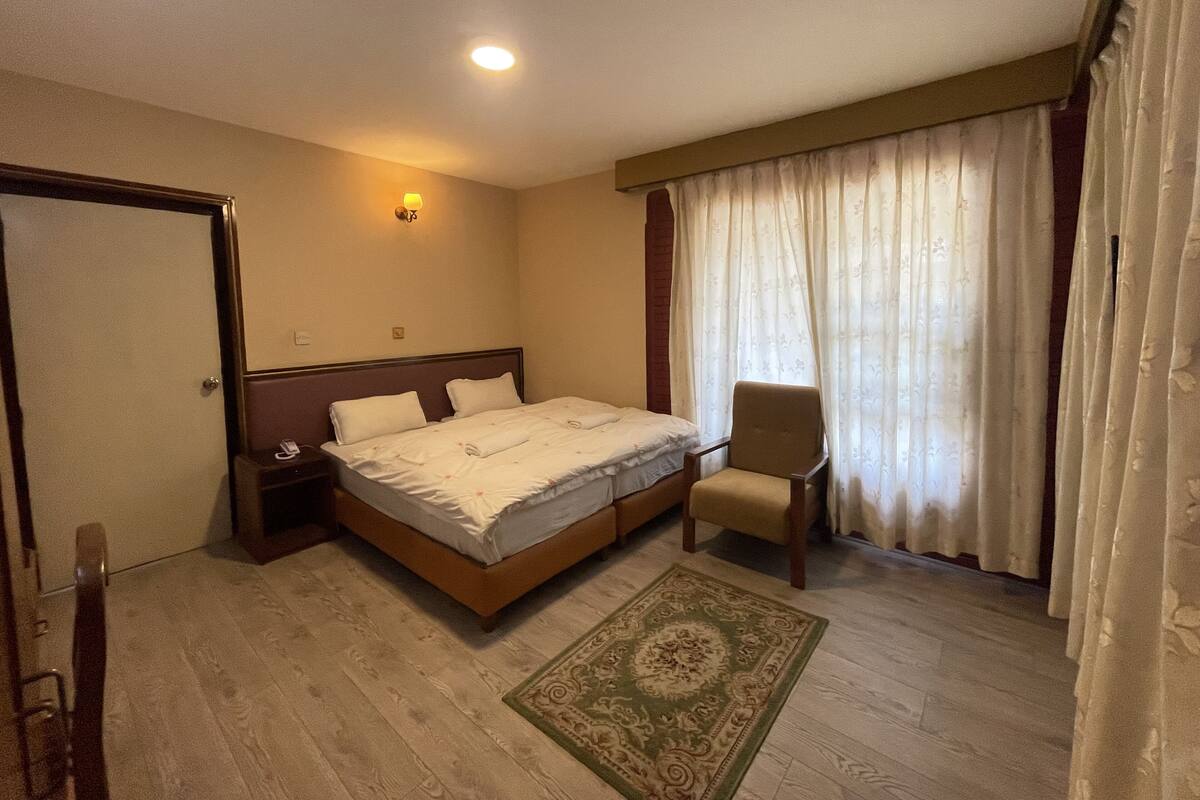 Habitación de hotel con cama, una silla, mesita de noche, lámpara y ventana con cortinas.