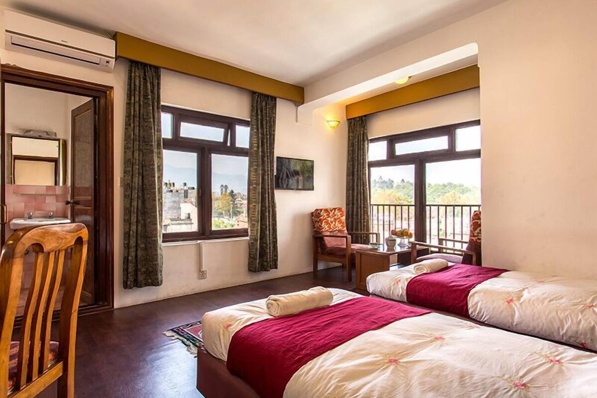 Habitación de hotel con dos camas, una silla de madera, una mesita con un florero y amplios ventanales con vistas al exterior.