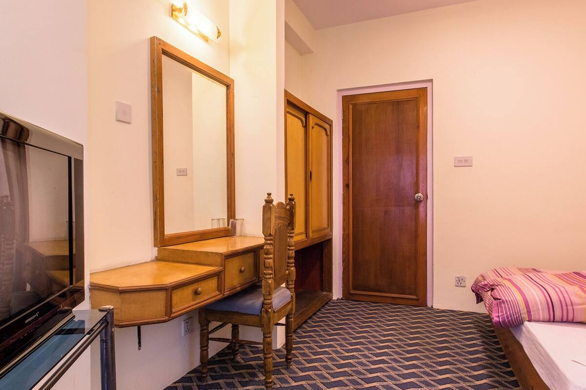 Una habitación pequeña y ordenada con un aparador de madera, un espejo, una cama con manta a rayas y un televisor sobre un mueble.