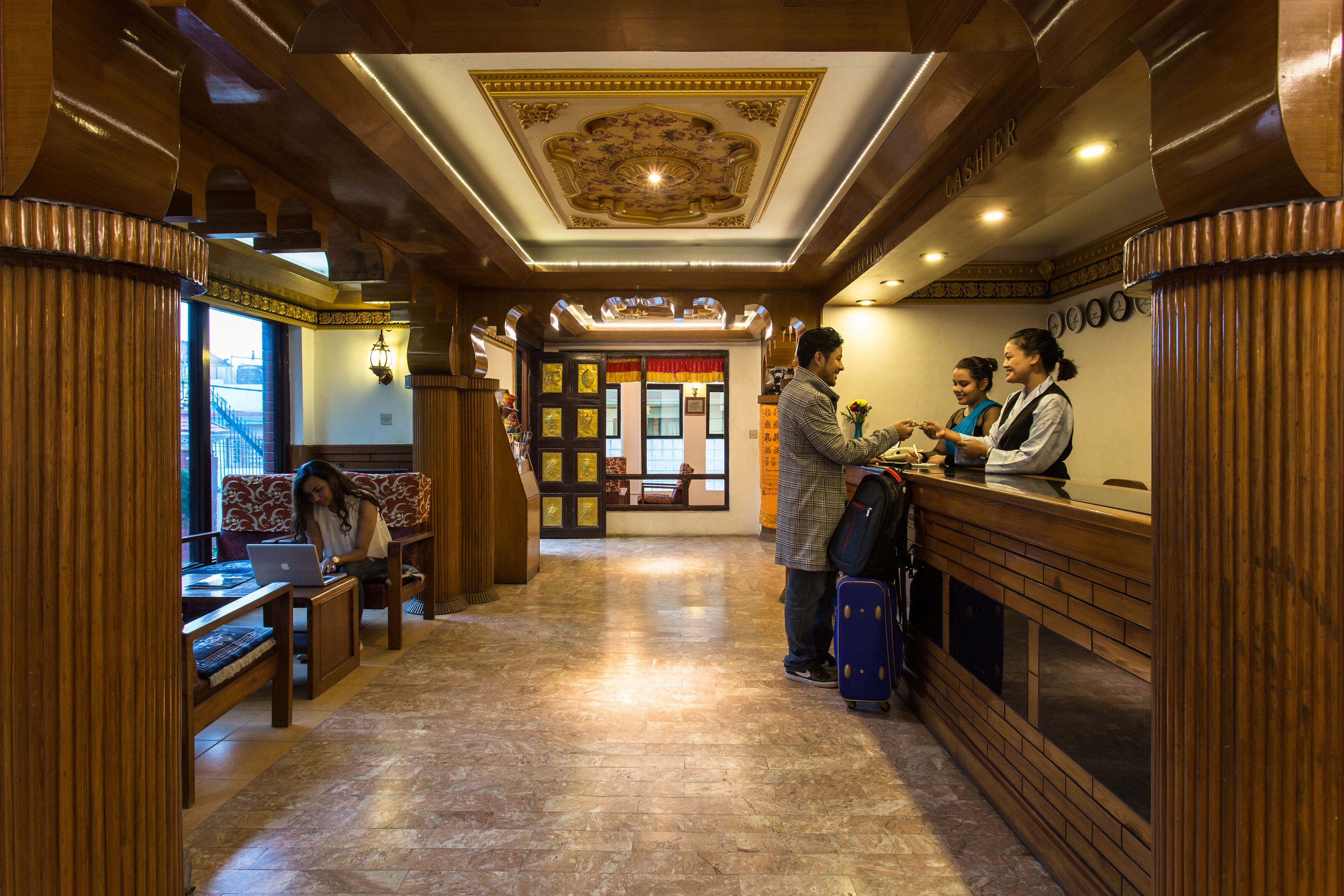 A hotel lobby with wooden columns, a reception desk, and a seating area with a person using a laptop.