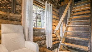A log cabin interior with a white leather chair, a wooden staircase, and a window with curtains.