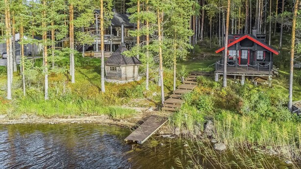 A cabin with a red roof, surrounded by tall trees and a small dock extending into a calm body of water.