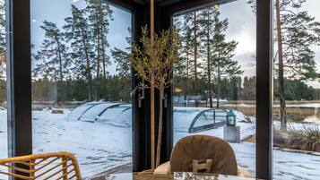 A room with a large window overlooking a snowy landscape, a table with a chair, and a vase with branches.