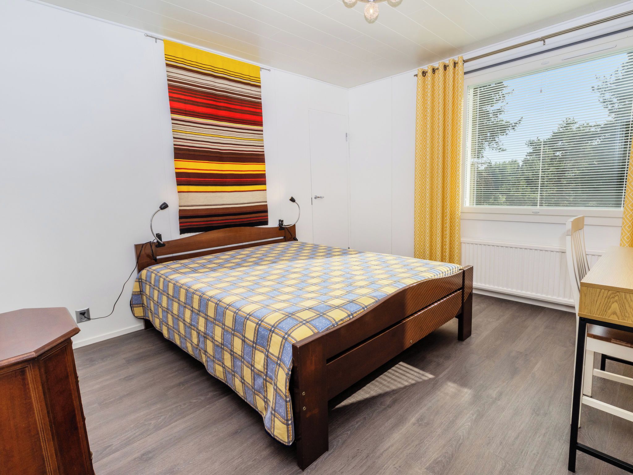 Ein Schlafzimmer mit einem hölzernen Bett, einem Schreibtisch, einem Stuhl, einem Fenster mit Jalousien und einem Streifenmuster-Vorhang.