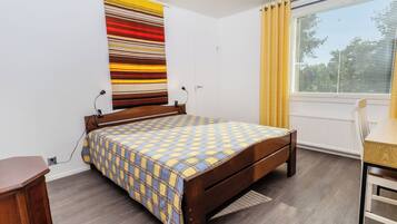 Ein Schlafzimmer mit einem hölzernen Bett, einem Schreibtisch, einem Stuhl, einem Fenster mit Jalousien und einem Streifenmuster-Vorhang.