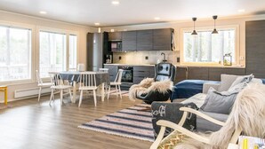 Ein modernes Wohnzimmer mit einer Couch, einem Esstisch mit Stühlen, einer Küchenzeile mit dunklen Schränken und großen Fenstern, die für natürliches Licht sorgen.