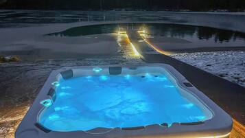 A hot tub with a wooden deck extending into a body of water, surrounded by a snowy landscape.