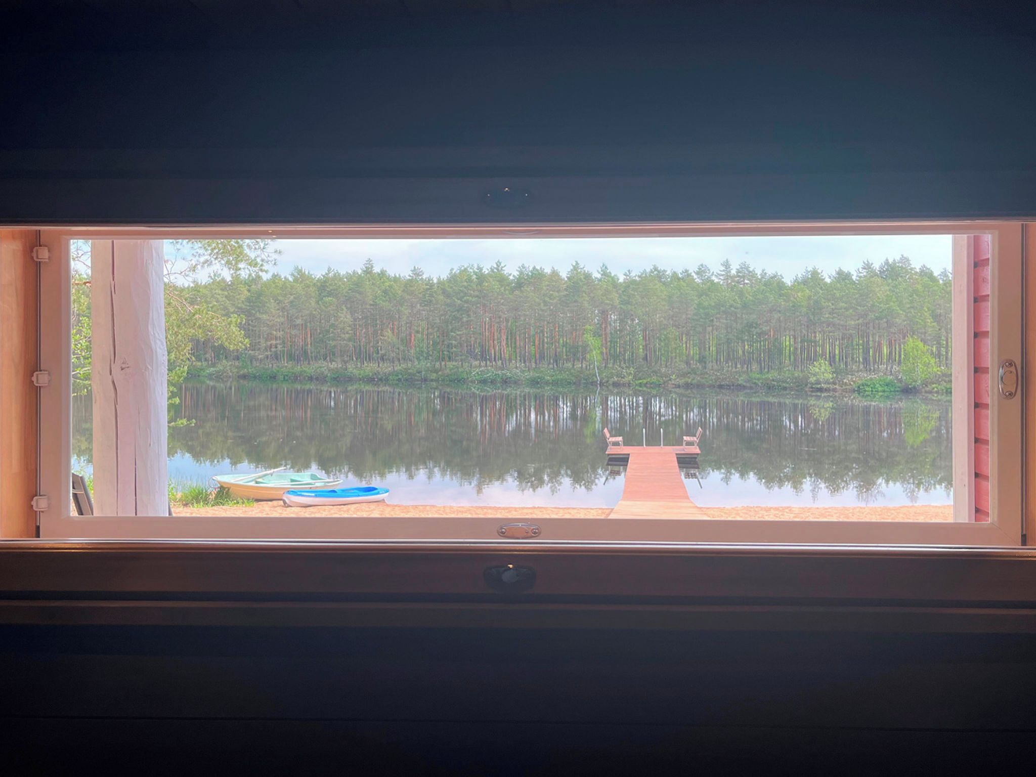 Ikkunasta avautuu näkymä tyyneen järveen, laituriin ja metsään.