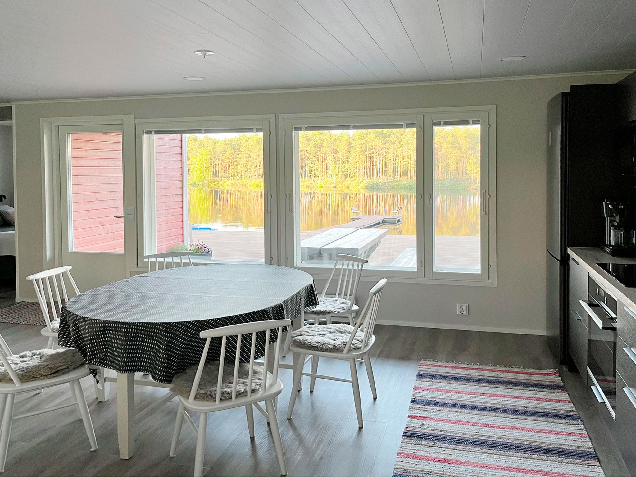 A dining area with a round table and chairs, large windows with a view of a wooded area, and a kitchen with modern appliances.