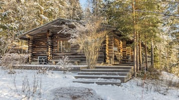 Une cabane en rondins dotée d’un porche et d’un escalier menant à l’entrée, entourée d’arbres couverts de neige.