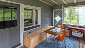 A cabin interior with a wooden bench, a table, two chairs, and a window overlooking a forest.
