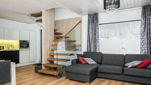 A modern living room with a grey sectional sofa, a wooden staircase, and a kitchen area with white cabinets and a black countertop.
