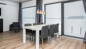 A modern dining area with a white table and grey chairs, large windows with blinds, and a ceiling with wooden panels.