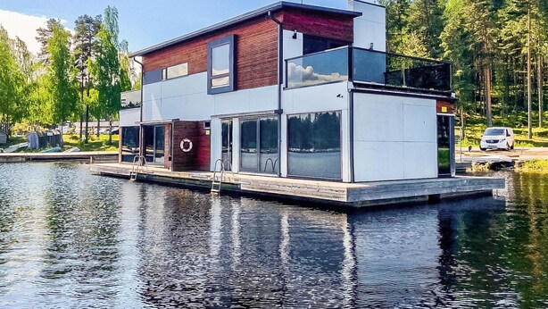 A modern houseboat with a flat roof, large windows, and a dock extending into a body of water.