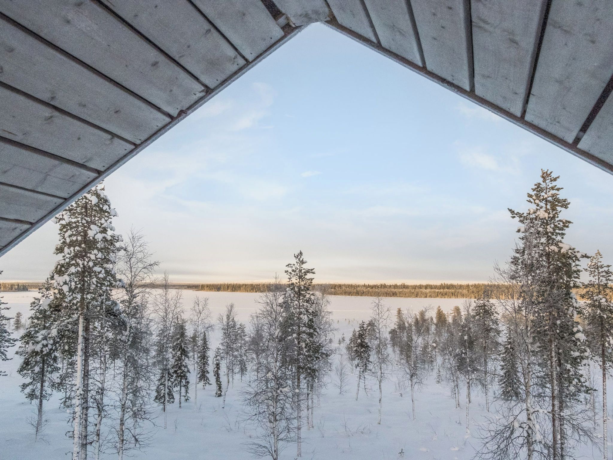 En vy genom ett triangulärt fönster med ett snötäckt landskap, inklusive träd och en frusen sjö eller flod.