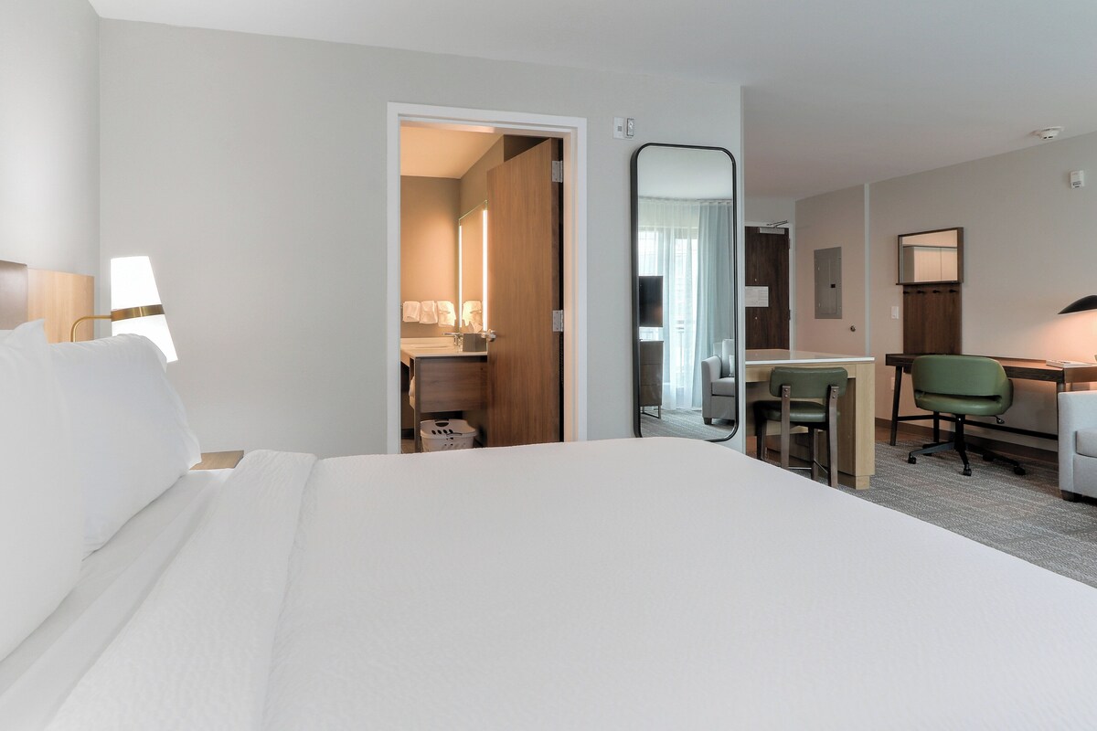 A hotel room with a bed, a desk, a chair, a mirror, and a bathroom visible through an open door.