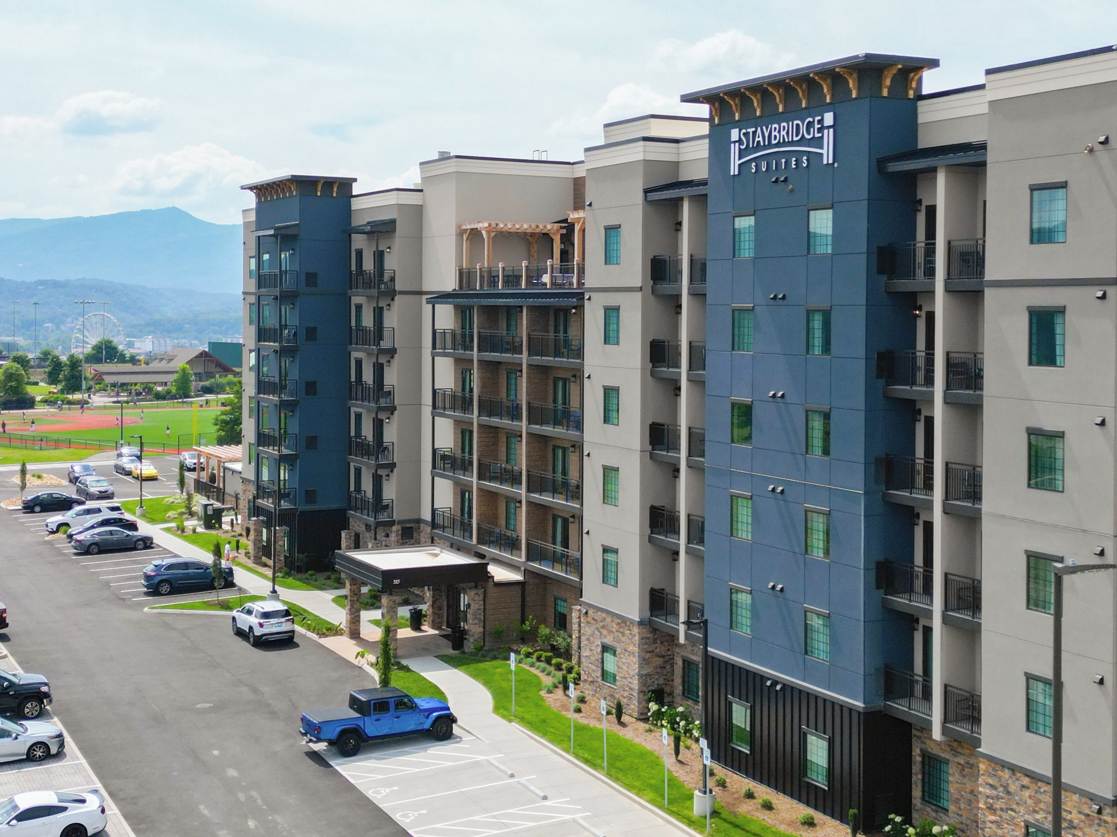 A multi-story hotel with Staybridge Suites branding, featuring a parking lot with several cars, a covered entrance, and a view of a baseball field in the background.