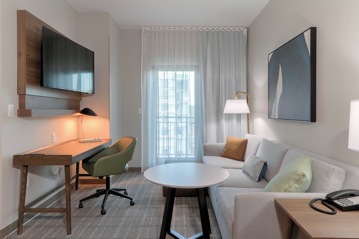 Habitación de hotel moderna con un escritorio de madera, una silla verde, un sofá blanco y un televisor de pantalla plana montado en la pared.