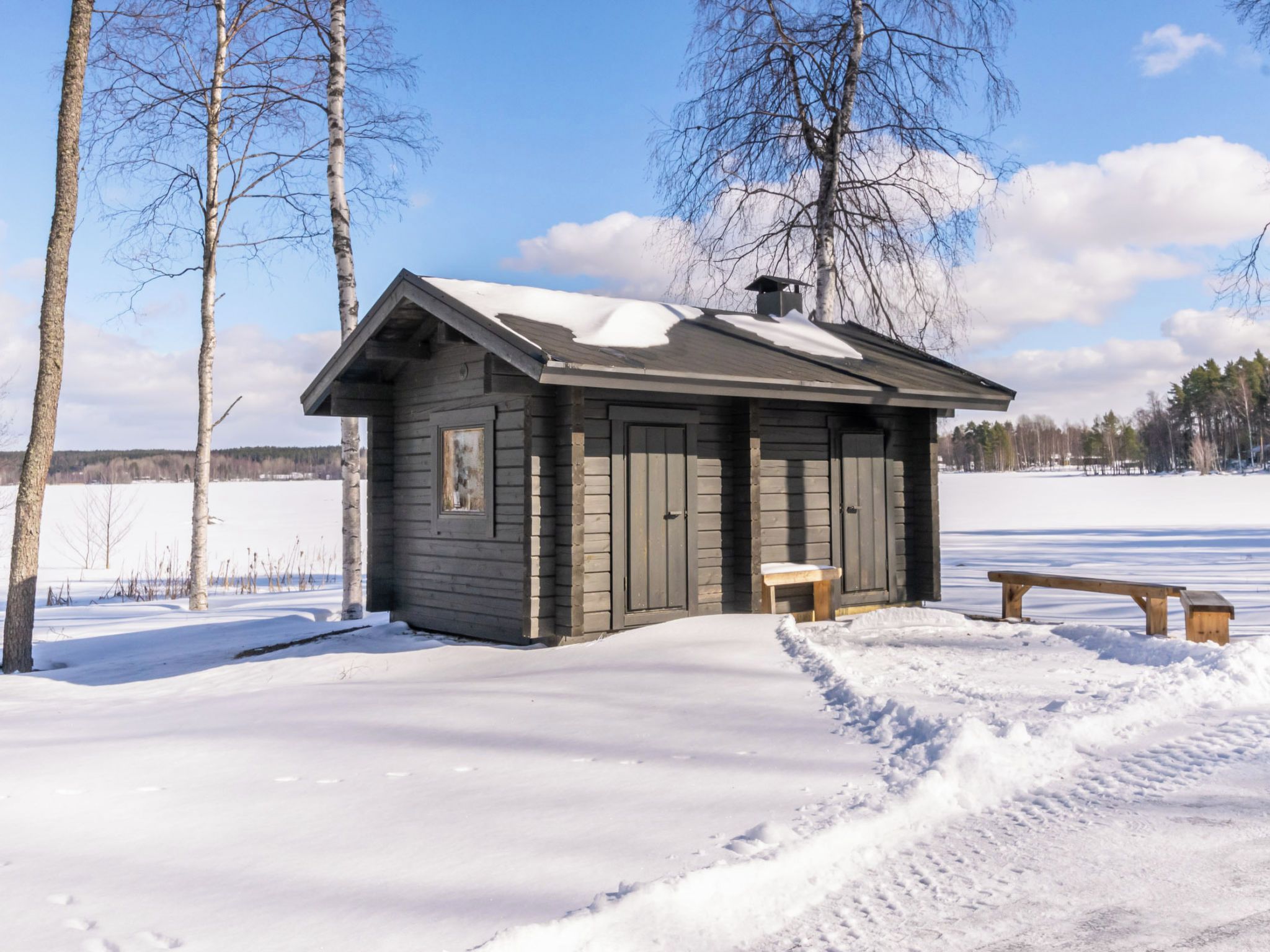 En stuga av trä med en bänk och ett snötäckt tak, omgiven av träd och ett snöigt landskap.