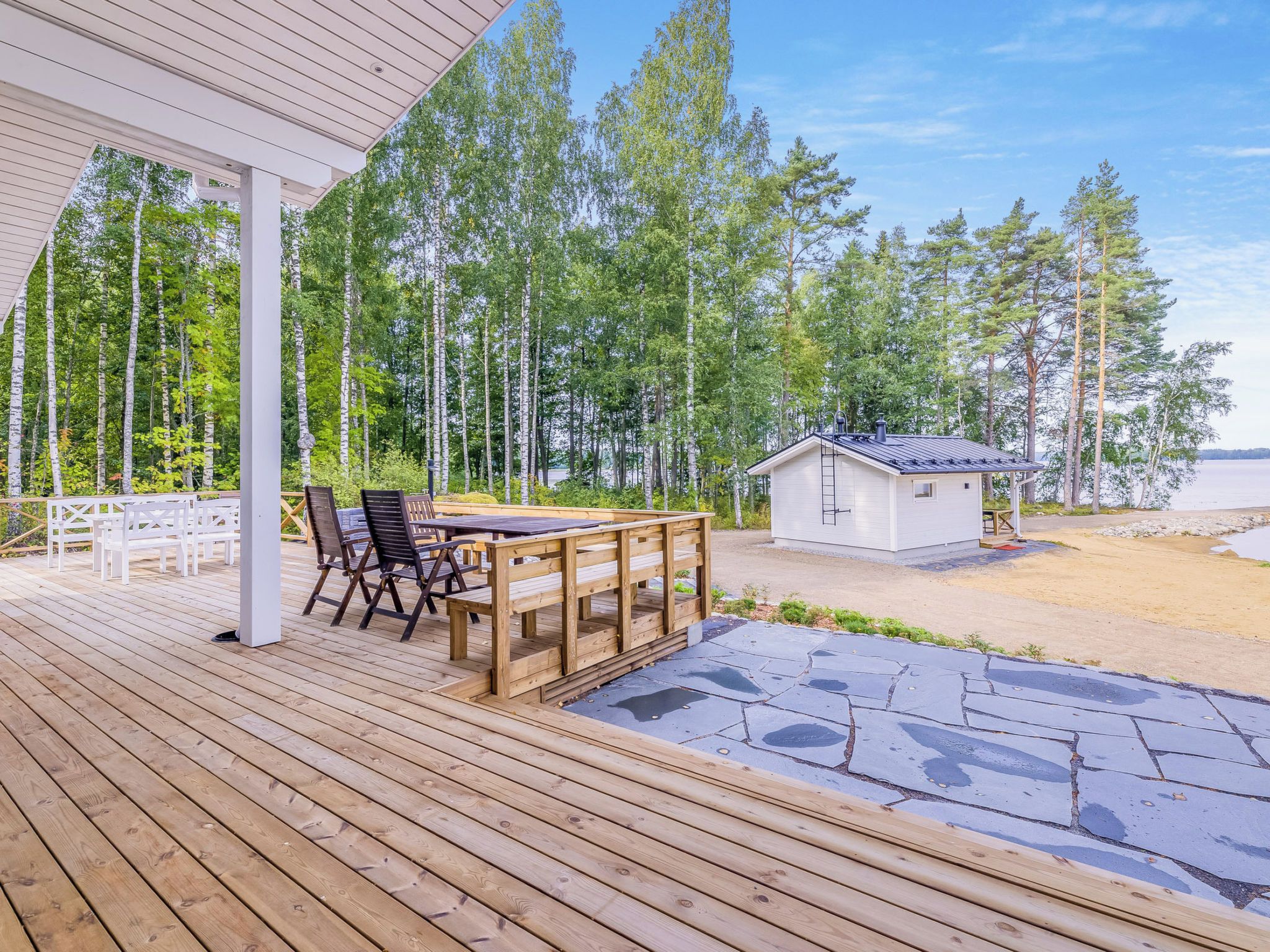 A wooden deck with a table and chairs, a small white building, and a forested area in the background.
