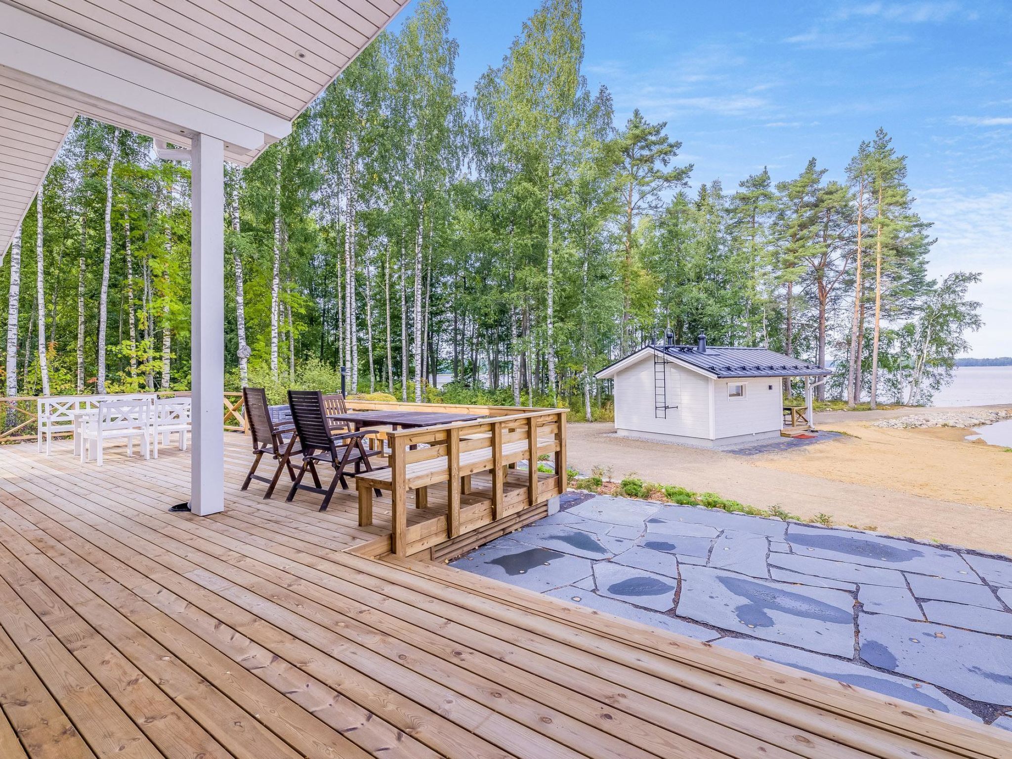 En træterrasse med et bord og stole, en mindre hytte, og et skovområde i baggrunden.