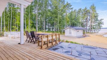 En træterrasse med et bord og stole, en mindre hytte, og et skovområde i baggrunden.