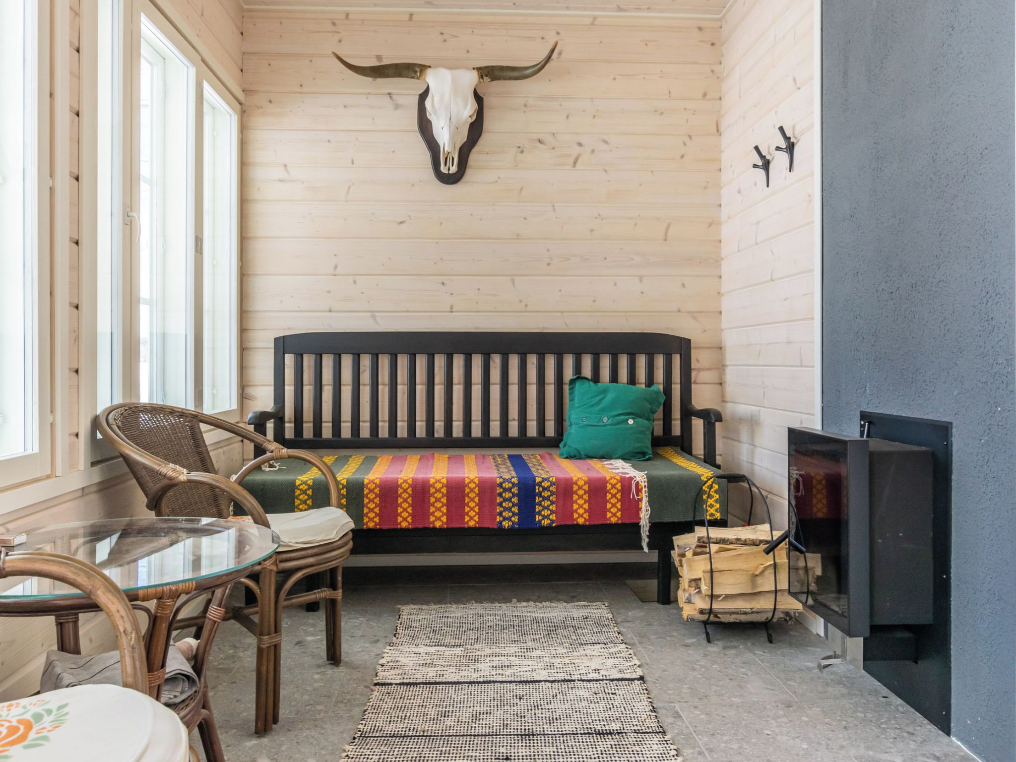 A cozy room with a wooden wall, a bed with a colorful blanket, a wicker chair, a glass table, and a fireplace.