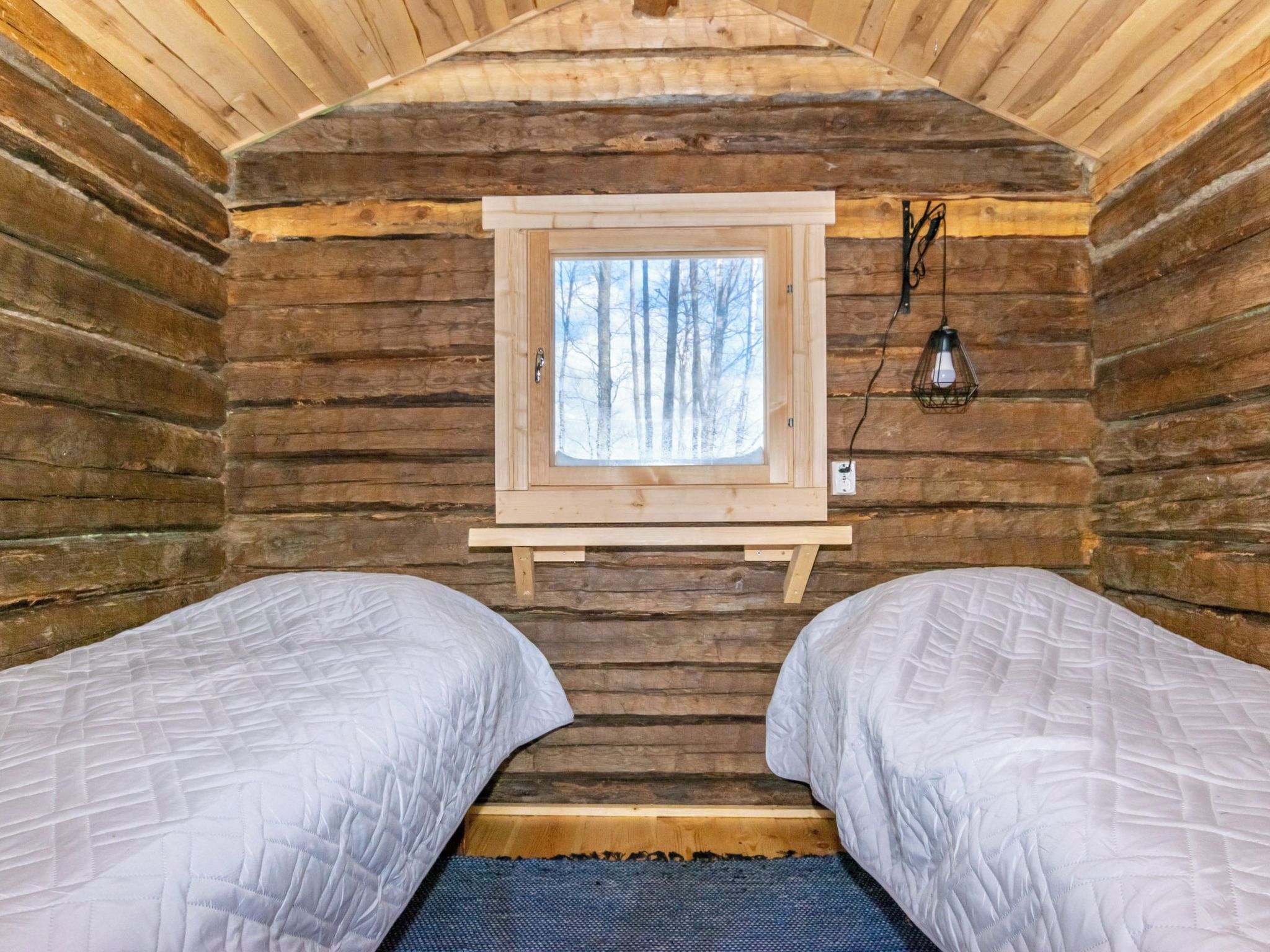 A cabin interior with two beds, a window with a view of trees, and a wall-mounted light fixture.