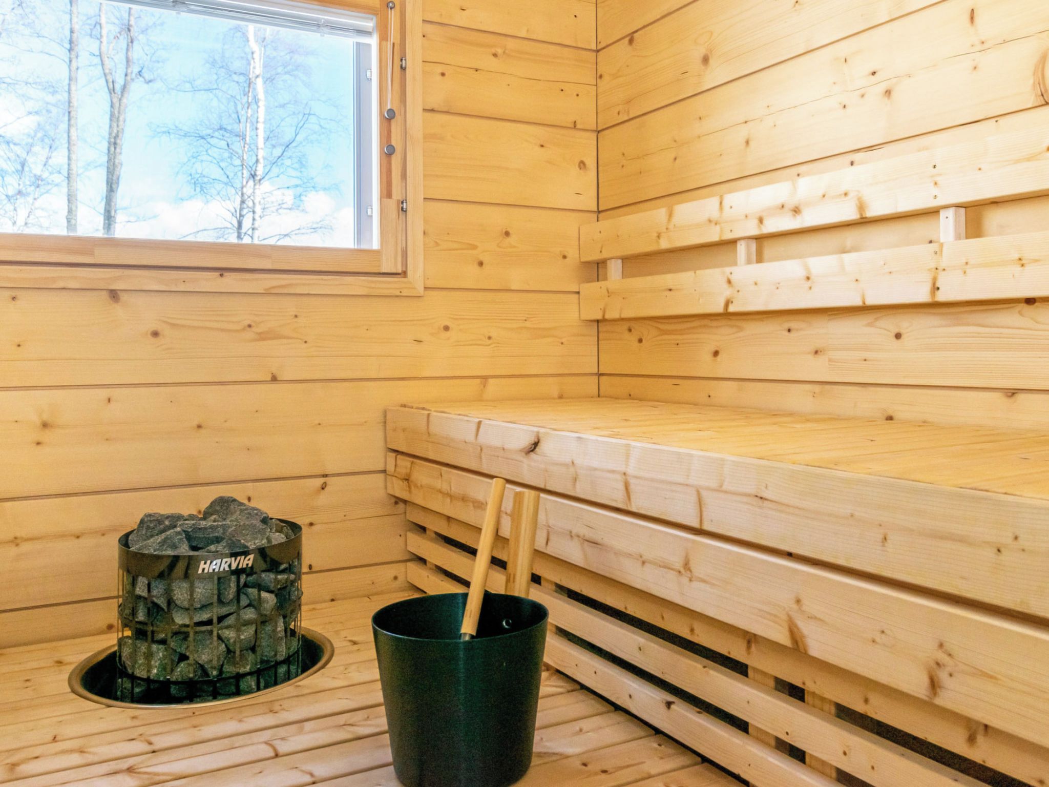 A wooden sauna room with a bench, a stone heater labeled 'HARVIA,' and a bucket with a stick.