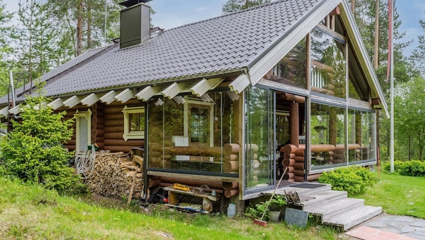 A log cabin with a glass extension, surrounded by trees and a grassy area.