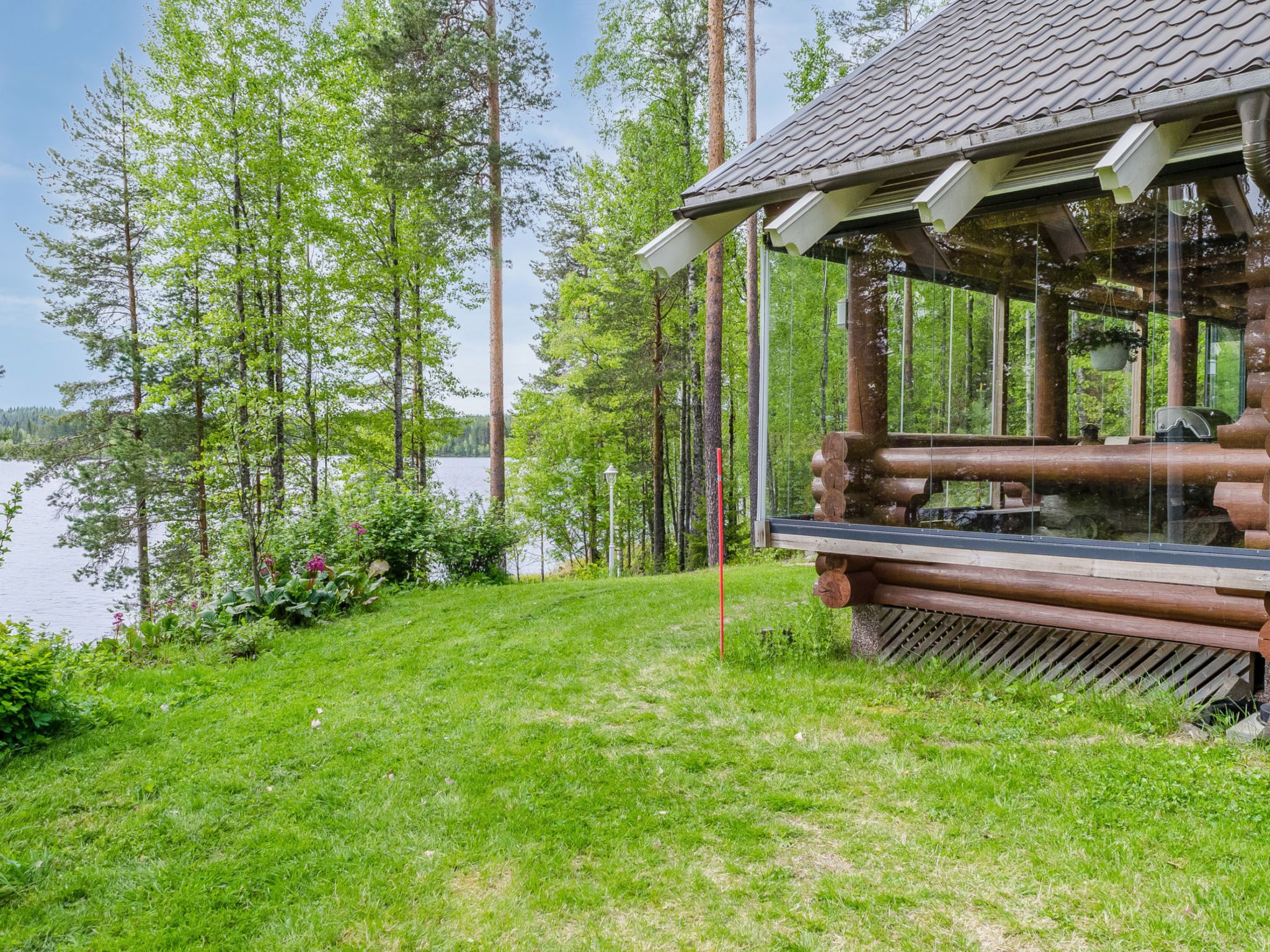 A wooden cabin with a glass enclosure, surrounded by a grassy area and trees, overlooking a body of water.
