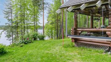 A wooden cabin with a glass enclosure, surrounded by a grassy area and trees, overlooking a body of water.