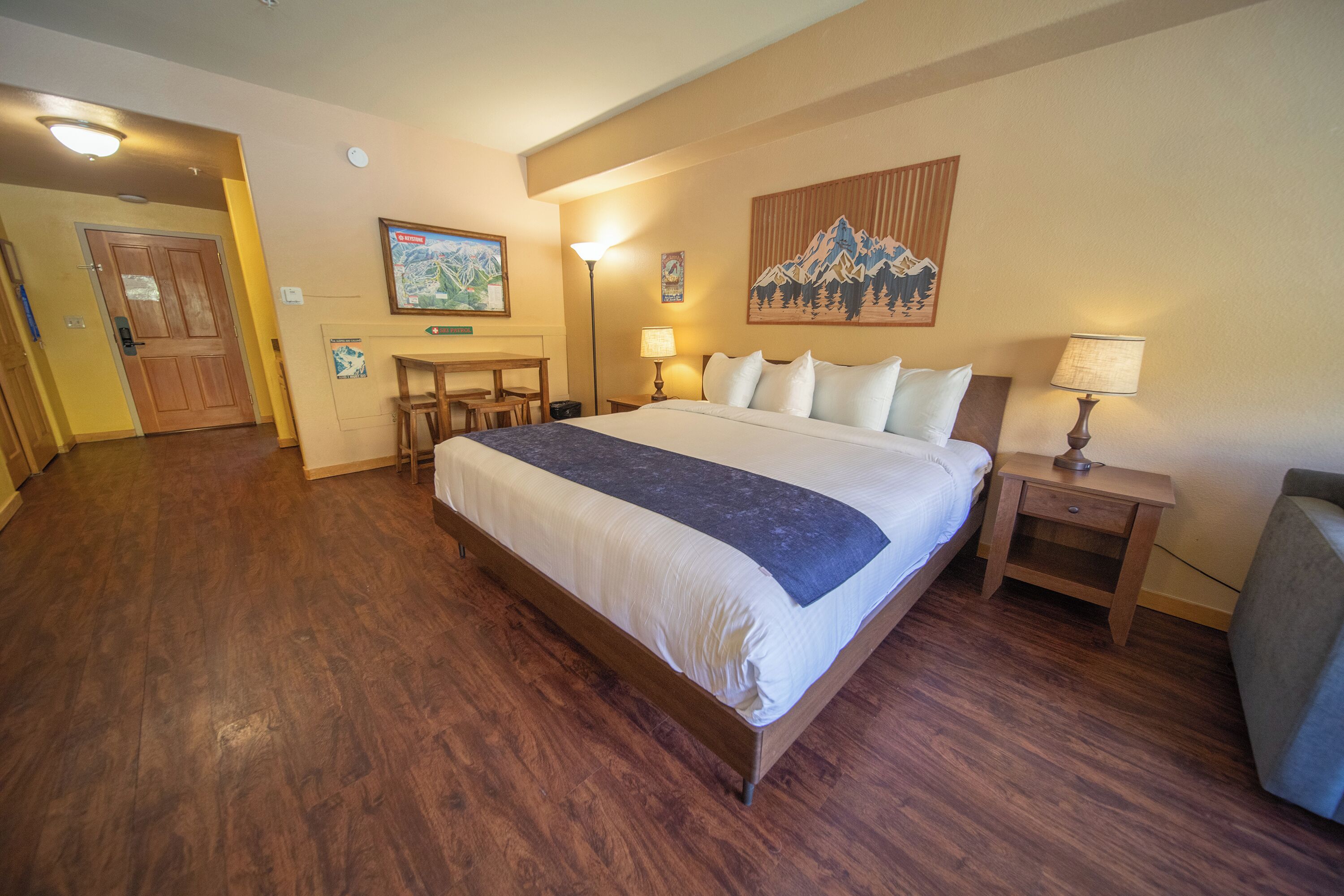 A hotel room with a large bed, wooden flooring, a desk with chairs, a lamp, and a painting on the wall.