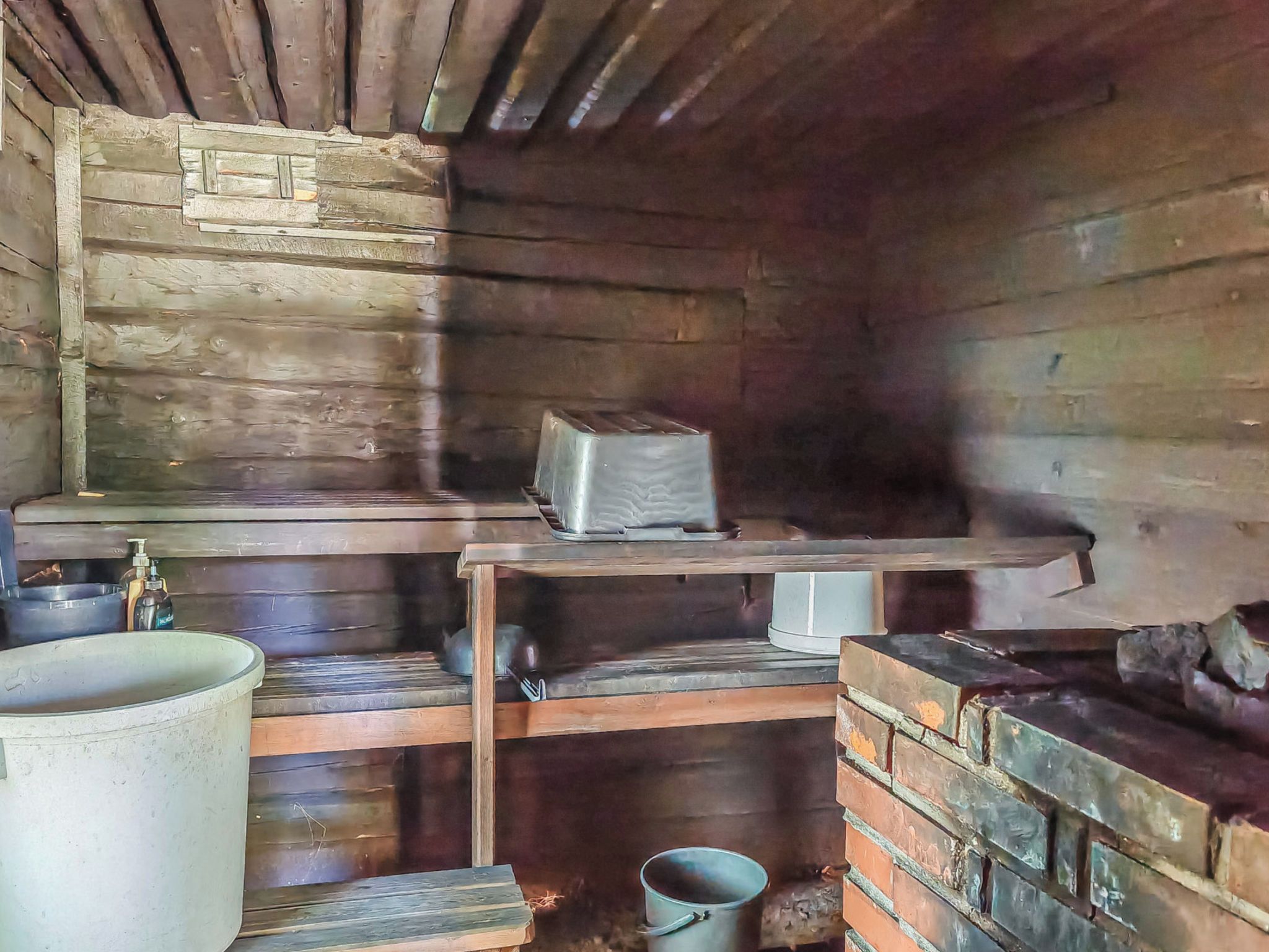 Interior rústico de una cabaña de madera con un horno de metal, un estante con una caja y un contenedor metálico grande.