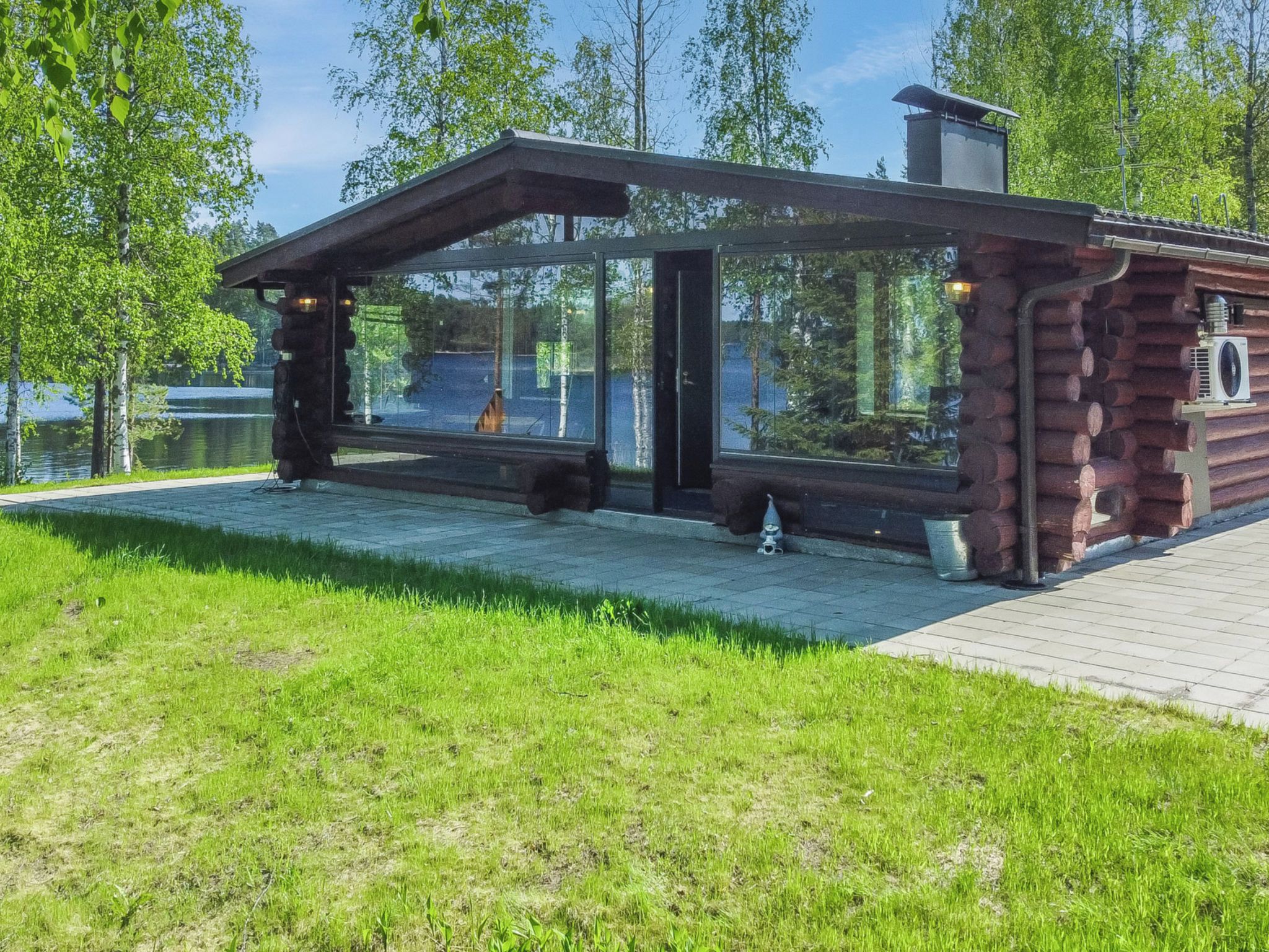 En modern timmerstuga med en glasveranda, belägen vid en sjö med en gräsklädd förgrund och träd i bakgrunden.