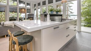 A modern kitchen with a large island, white cabinetry, and large windows offering views of greenery.