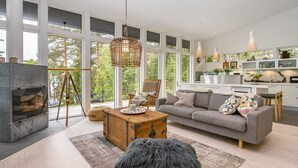 A modern living room with a fireplace, sofa, coffee table, and large windows offering a view of greenery.