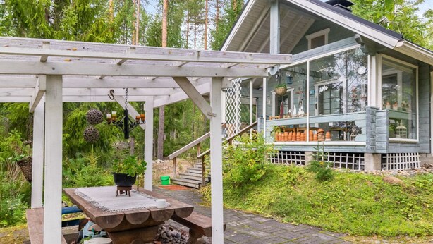 Una pérgola de madera con una mesa y una lámpara colgante, que conduce a una casa con un porche y plantas en macetas.