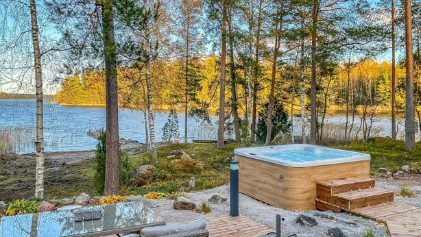 A wooden deck with a hot tub, a glass table, and a wicker sofa overlooking a lake surrounded by trees. - Villa havet by Interhome (Porvoo)