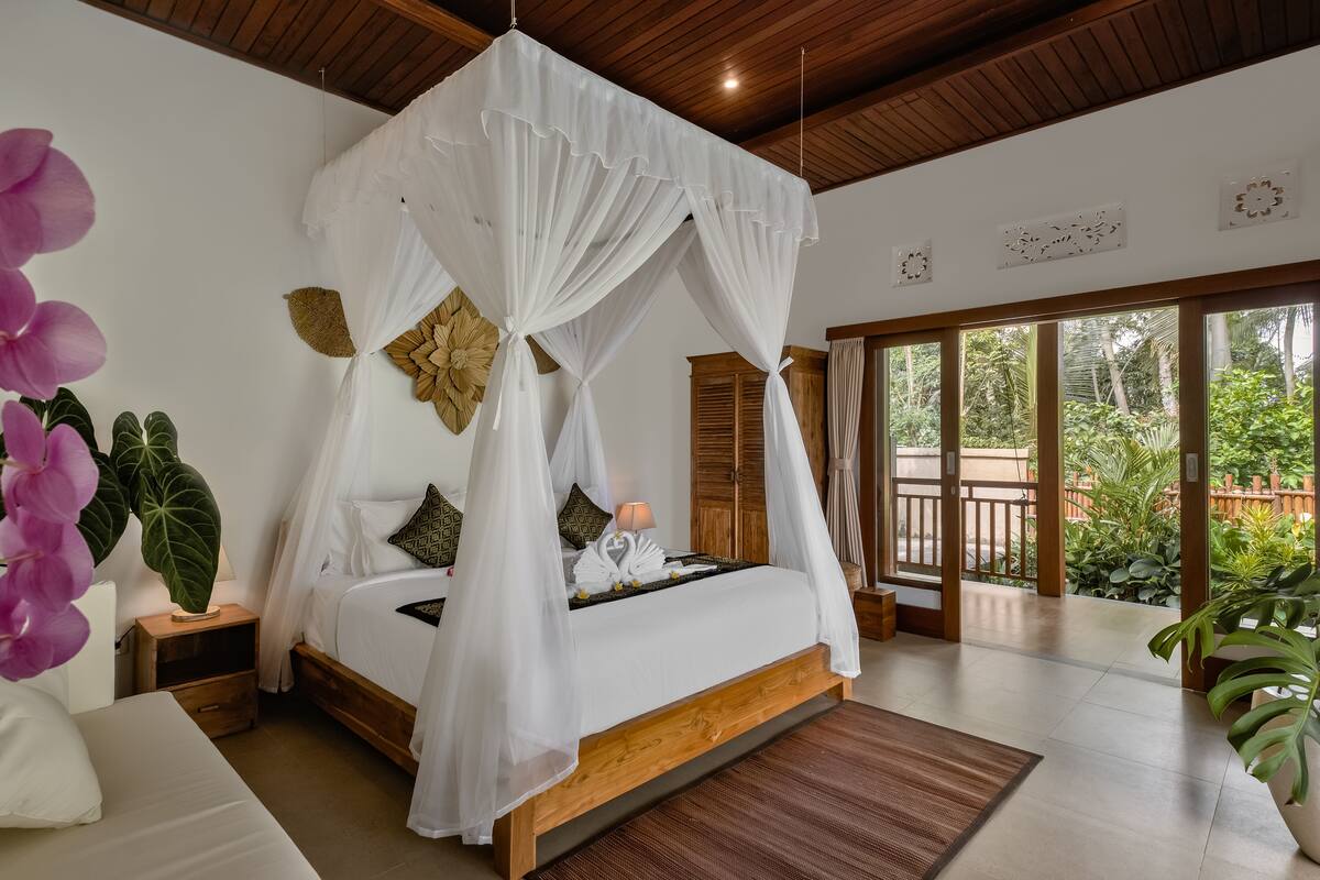 A bedroom with a canopy bed, wooden headboard, bedside tables, and a view of greenery through large windows.