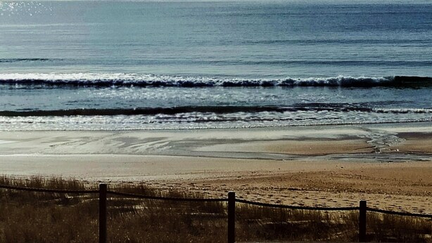 Una platja amb una tanca al primer pla, mar tranquil·la i terra a la distància amb un veler.