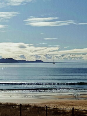 A beach with a fence in the foreground, calm sea, and distant land with a sailboat. - Family Beach Bach (Opito Bay)