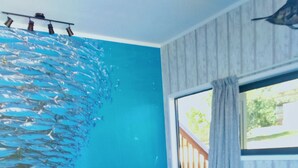 Un sofà seccional en forma de L amb un coixí, una paret blava amb un mural de peixos, i una finestra amb vista a vegetació.