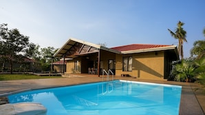 A swimming pool with a covered patio area, a house with a red roof, and palm trees in the background.
