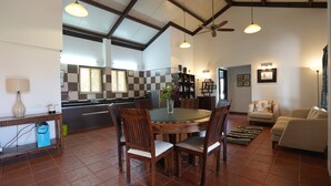 A spacious living area with a round dining table surrounded by wooden chairs, a sofa, and a coffee table. A kitchen with a checkered backsplash and modern appliances is visible in the background.