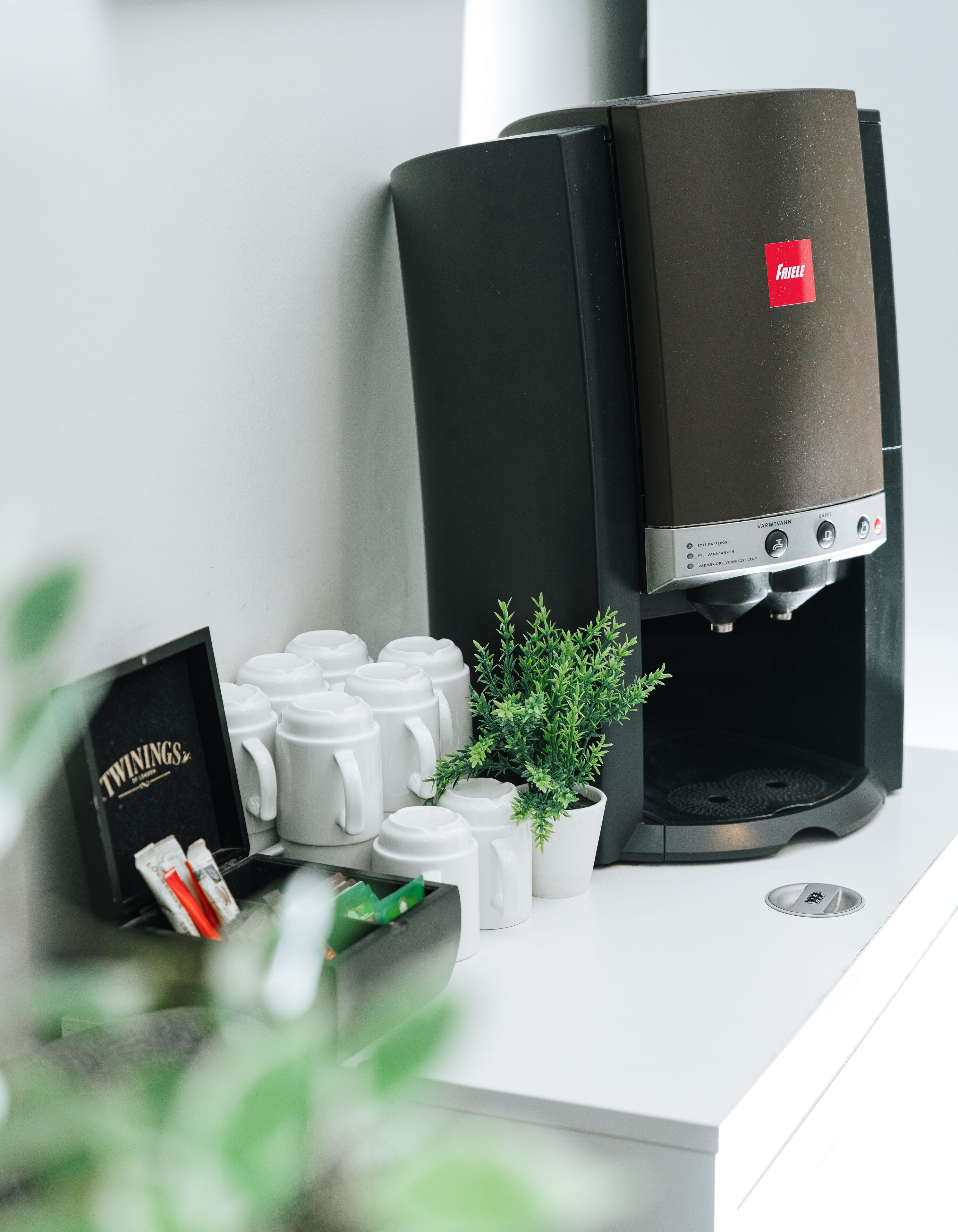 Una cafetera con etiqueta roja, una planta en maceta y un portavasos con tazas blancas y una caja de 'Twinings'.