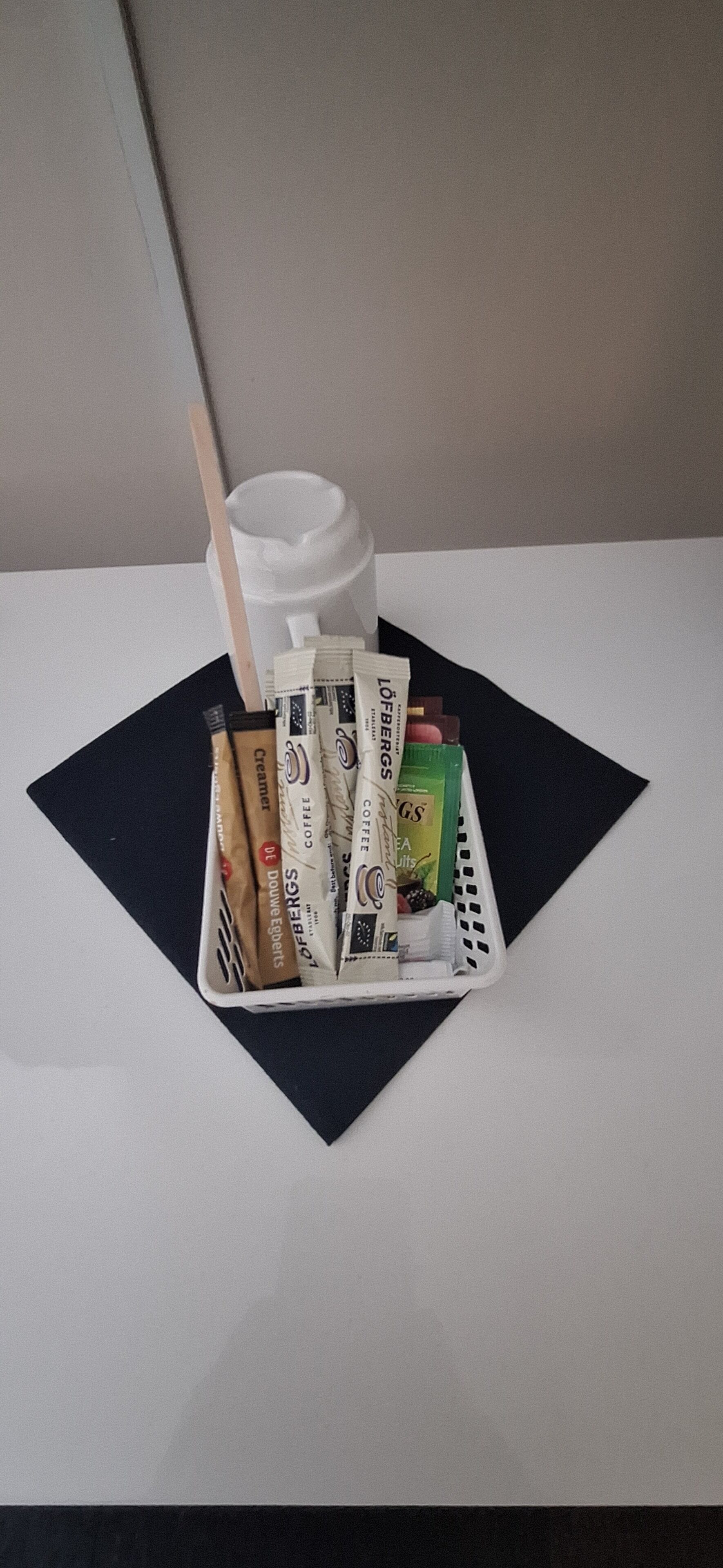 Una tazza di caffè bianca con un cucchiaino di legno su un sottopiatti nero. Un cesto bianco contiene bustine di caffè e una bottiglia verde.