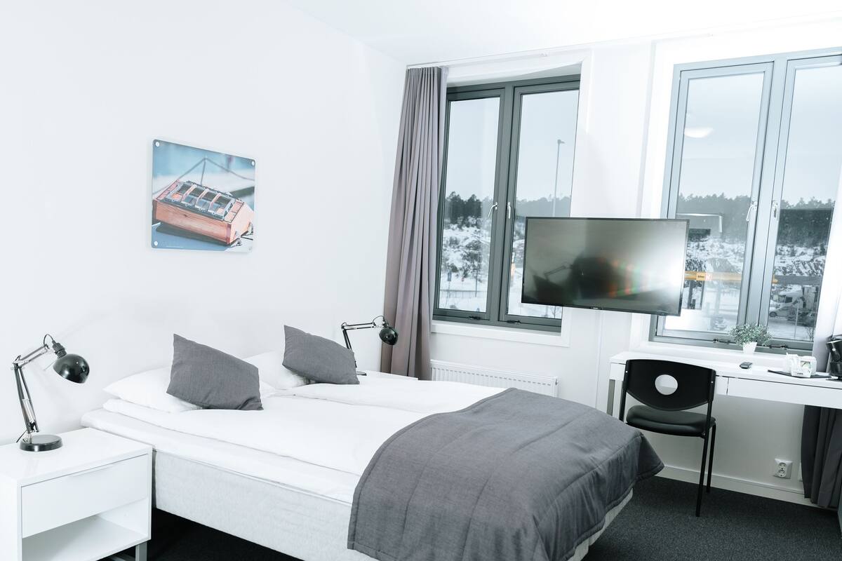 A modern hotel room with a bed, bedside lamps, a desk with a chair, a TV, and a window with a view of a snowy landscape.