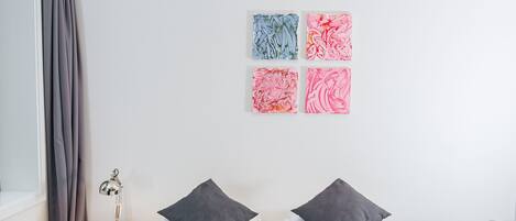 A modern hotel room with a neatly made bed, a bedside table with a lamp, and a wall with four abstract paintings.