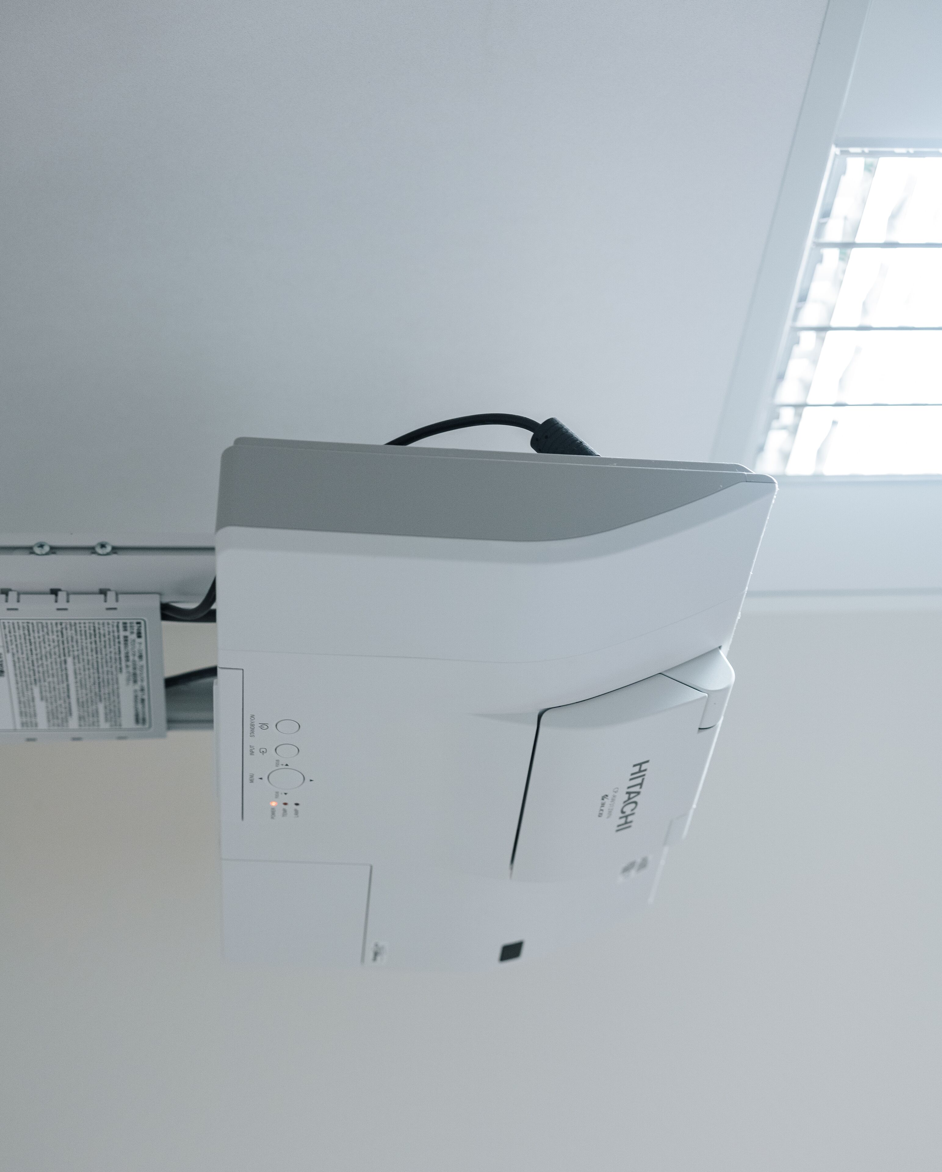 En hvid printer med sort kabel, placeret i et rum med et vindue og en vægmonteret aircondition.
